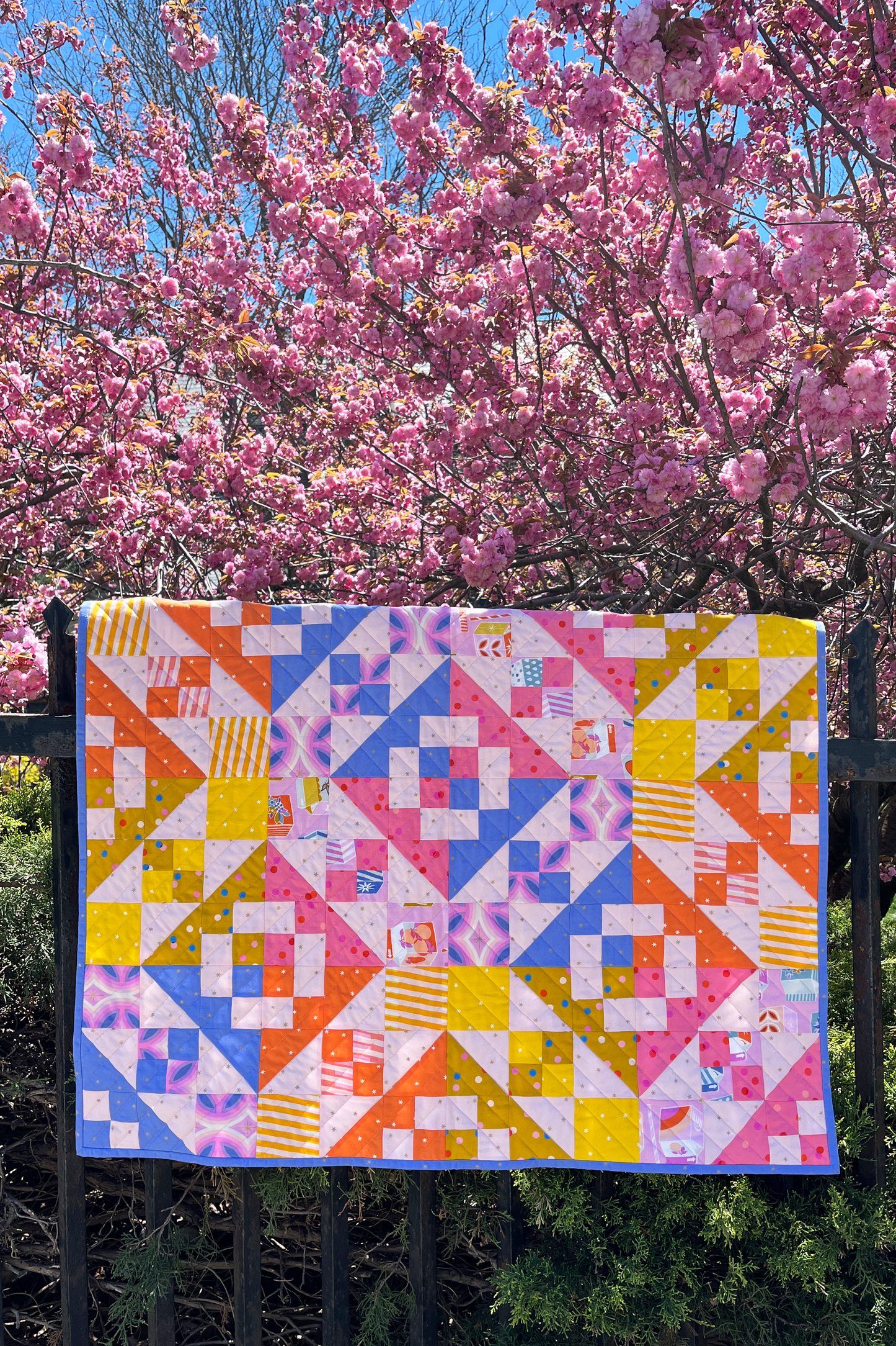 Patchwork quilt hanging on a fence against a backdrop of pink cherry blossom trees