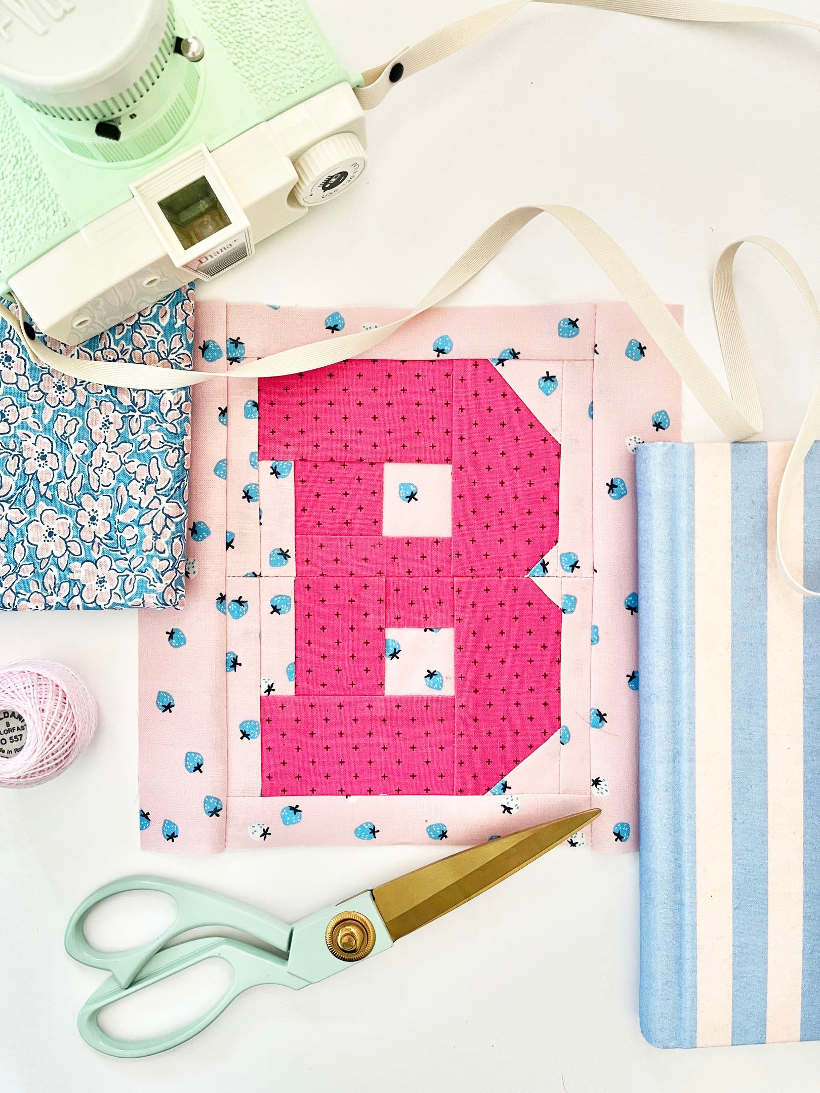 Varsity letter B quilt block in hot pink on powder pink, surrounded by desktop accessories like a mint green camera, striped notebook, mint green scissors, and floral fabric.